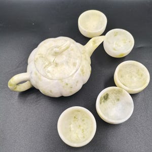 Könnte beinhalten: Ein Teeservice aus blassgrünem und weißem, gesprenkeltem Stein, bestehend aus einer Teekanne mit Deckel und vier kleinen passenden Teetassen. Die Teekanne hat einen abgerundeten, segmentierten Körper mit einem gebogenen Ausguss und einem Griff.