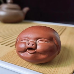 Może przedstawiać: Mała, czerwonobrązowa ceramiczna figurka uśmiechniętej twarzy z zamkniętymi oczami i nosem w kształcie świni. Figurka jest okrągła i gładka, ze szczegółowymi rysami twarzy. Czajnik jest rozmyty w tle.
