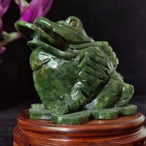 May include: A dark green jade frog sculpture, holding a coin in its mouth, sits atop a wooden stand. The frog is detailed with carved features and sits on a base of carved leaves. The sculpture is approximately 13 cm long.