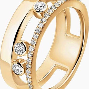 Könnte beinhalten: Ein goldfarbener Ring mit drei runden, klaren Edelsteinen und einem Band aus kleinen, klaren Edelsteinen. Der Ring hat ein einzigartiges, offenes Design mit einem Doppelband und einem dekorativen Band aus kleinen Edelsteinen.