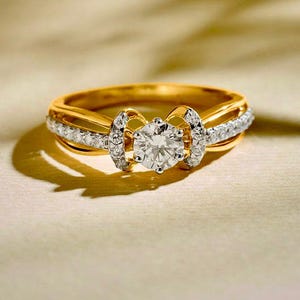 Peut inclure: Une bague en or avec une pierre centrale ronde et transparente montée sur griffes, entourée de deux éléments décoratifs. L'anneau est divisé en deux sections parallèles, chacune incrustée d'une rangée de petites pierres transparentes.