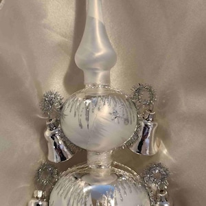 May include: A decorative Christmas tree topper with a pearlescent finish. The topper features two frosted glass spheres with silver glitter accents and small silver bells. The top has a long, slender, pointed tip.
