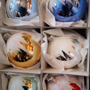 Op de afbeelding: Zes glazen kerstballen in een doos. De ballen zijn rond en hebben een witte, frosted afwerking met een zilveren glitter-effect. De ballen zijn in de kleuren blauw, goud, wit, groen en rood.