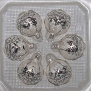 Op de afbeelding: Zes zilveren en witte kerstballen met een geometrisch ontwerp in een witte plastic container.