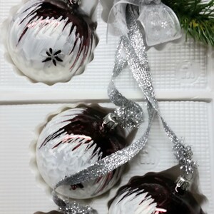 Op de afbeelding: Vijf witte en bordeauxrode kerstballen met een zilveren glitterafwerking en een sneeuwvlokkenpatroon. De ballen bevinden zich in een witte doos met een zilveren lint en een zilveren strik.