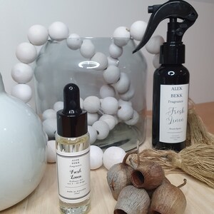 May include: Two bottles of home fragrance oil, one in a clear glass bottle with a black dropper and the other in a black bottle with a black spray nozzle. Both bottles have white labels with black text that reads "Fresh Linen" and "Alex Bekk Fragrance". The bottles are sitting on a wooden surface with a string of white beads and dried eucalyptus pods.