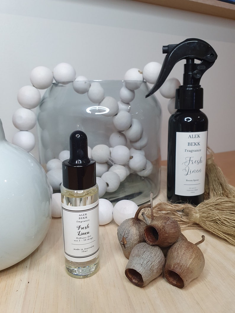 May include: Two bottles of home fragrance oil, one in a clear glass bottle with a black dropper and the other in a black bottle with a black spray nozzle. Both bottles have white labels with black text that reads "Fresh Linen" and "Alex Bekk Fragrance". The bottles are sitting on a wooden surface with a string of white beads and dried eucalyptus pods.