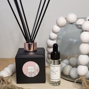 May include: Black diffuser bottle with a pink and white marble sticker with the letters AB on it. The bottle has black diffuser sticks and a rose gold cap. A clear glass bottle with a black cap and a white label that says "Lychee and Peony Diffuser Oil" is next to the diffuser bottle.