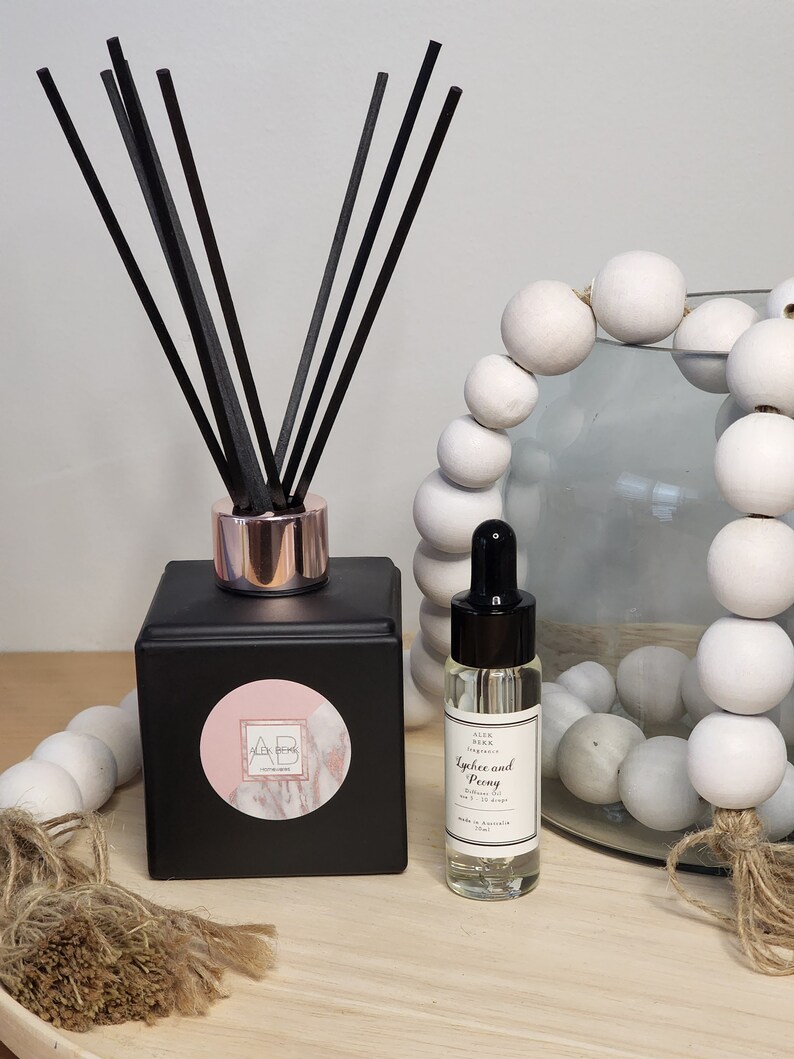May include: Black diffuser bottle with a pink and white marble sticker with the letters AB on it. The bottle has black diffuser sticks and a rose gold cap. A clear glass bottle with a black cap and a white label that says "Lychee and Peony Diffuser Oil" is next to the diffuser bottle.