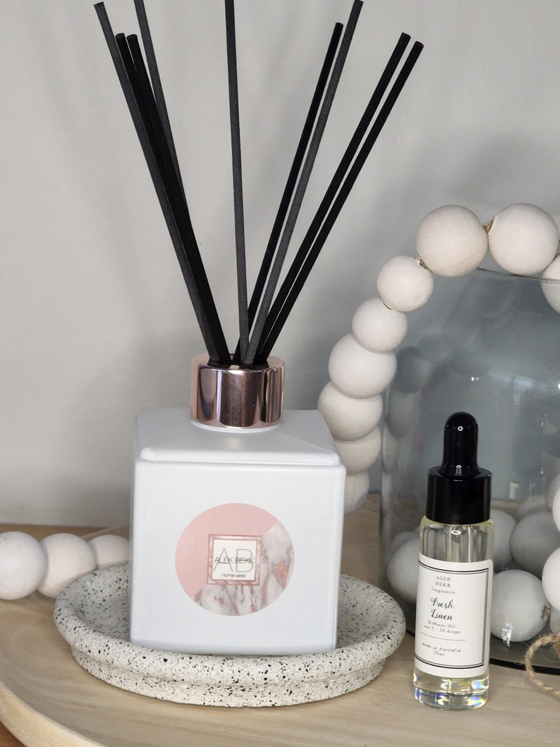 May include: White diffuser bottle with black reeds and a pink and white marble design label. The label says "Alex Beek Home Fragrance". A small bottle of essential oil with a black dropper and a white label that says "Alex Beek Fragrance Oil Fresh Linen" is also pictured.