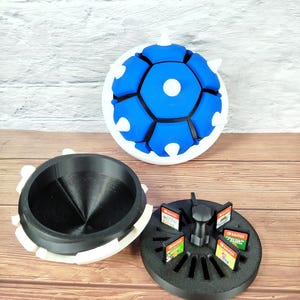 Puede incluir: Un soporte para cartuchos de juego con forma de caparazón azul con exterior blanco e interior azul. El soporte está abierto, revelando una base negra con cartuchos de juego. También se ve un contenedor separado en blanco y negro.