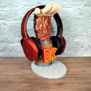 Könnte beinhalten: Ein rotes Headset liegt auf einem 3D-gedruckten Kopfhörerständer. Der Ständer hat ein braunes Fass, eine rote, ananasförmige Oberseite mit einer Hand und eine weiße Basis. Die Buchstaben "DK" werden in Gelb und Rot angezeigt.