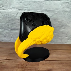 Puede incluir: Un mando de videojuegos negro descansa en un soporte amarillo y negro. El soporte tiene un brazo curvo que sujeta el mando y una base redonda. El mando es negro con botones y una cruceta direccional.