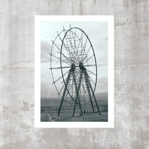 May include: A black and white photographic print featuring a large, skeletal metal ferris wheel structure standing in an open field. The wheel has a complex lattice of spokes and supports, set against a cloudy sky and a distant, low-lying landscape.