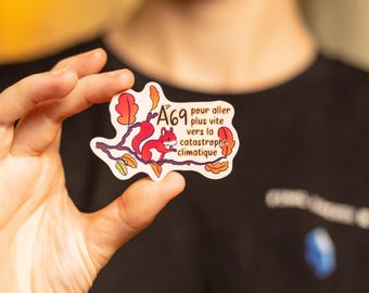 Op de afbeelding: Een sticker met een rood eekhoorntje op een tak met herfstbladeren. De sticker heeft de witte tekst "A69 pour aller plus vite vers la catastrophe climatique". De sticker wordt in een hand gehouden.