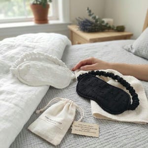 May include: A white and a black sleep mask with elastic bands, a small drawstring bag, and a tag with text on a bed. The white mask has a ruffled edge. The black mask is on a white cloth. The bag has the text "PURE MUSLIN SLEEP".