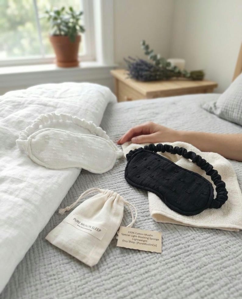 May include: A white and a black sleep mask with elastic bands, a small drawstring bag, and a tag with text on a bed. The white mask has a ruffled edge. The black mask is on a white cloth. The bag has the text "PURE MUSLIN SLEEP".
