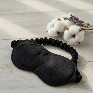 May include: A black sleep mask with a textured surface and elastic band. The mask is designed to block out light. Cotton and lavender sprigs are in the background.