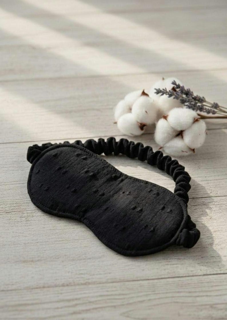 May include: A black sleep mask with a textured surface and elastic band. The mask is designed to block out light. Cotton and lavender sprigs are in the background.