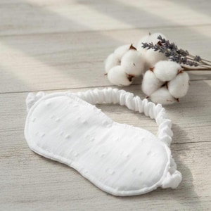 May include: A white eye mask with a textured surface and an elastic band, designed for sleep. The mask is accompanied by a sprig of lavender and cotton buds, suggesting a relaxing theme. The eye mask is made of soft material.