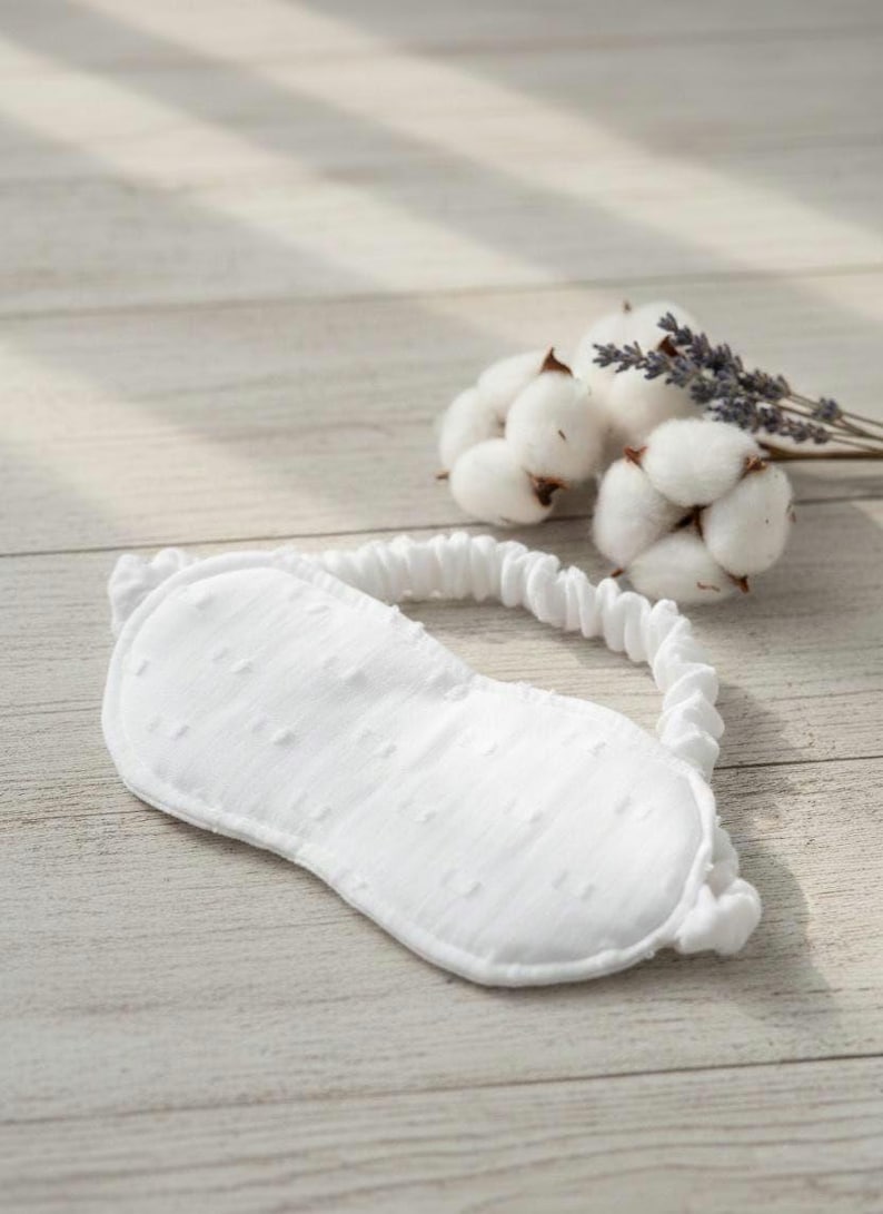 May include: A white eye mask with a textured surface and an elastic band, designed for sleep. The mask is accompanied by a sprig of lavender and cotton buds, suggesting a relaxing theme. The eye mask is made of soft material.