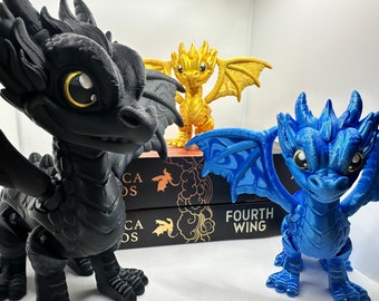 May include: Three small dragon figurines, one black, one gold, and one blue, are posed on a book titled "Fourth Wing".