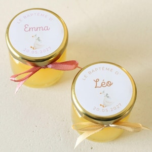 Peut inclure: Deux petits pots de miel en verre, avec des couvercles dorés et des étiquettes personnalisées. Les étiquettes indiquent "Emma" et "Léo" avec une date et une illustration. Chaque pot est orné d'un ruban.
