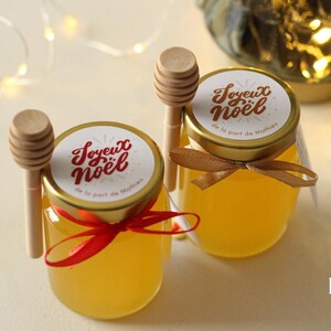Peut inclure: Deux petits pots en verre remplis de miel doré, chacun coiffé d'un couvercle et d'une étiquette « Joyeux Noël ». Des cuillères à miel en bois et des rubans décoratifs sont attachés. Les pots font partie d'un coffret cadeau de vacances.