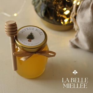 Peut inclure: Un petit pot en verre rempli de miel doré, surmonté d'un couvercle doré et d'une étiquette indiquant "JOYEUX NOEL". Un dipper à miel en bois repose à côté du pot, attaché avec un ruban marron. Le pot est étiqueté "LA BELLE MIELLEE".