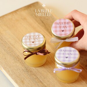 Peut inclure: Trois petits pots en verre avec des couvercles dorés et des étiquettes en vichy colorées. Les étiquettes indiquent "Agathe 1 an", "Hugo 1 an" et "Ambre 1 an". Les pots sont sur une surface en bois.