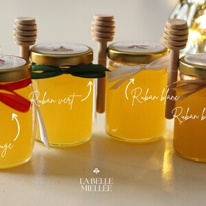 May include: Four small glass jars filled with golden honey, each with a gold lid and a decorative ribbon in red, green, white, and bronze. Wooden honey dippers rest beside each jar. The jars are labeled with the text "La Belle Miell&eacute;e".