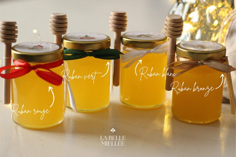 May include: Four small glass jars filled with golden honey, each with a gold lid and a decorative ribbon in red, green, white, and bronze. Wooden honey dippers rest beside each jar. The jars are labeled with the text "La Belle Miell&eacute;e".