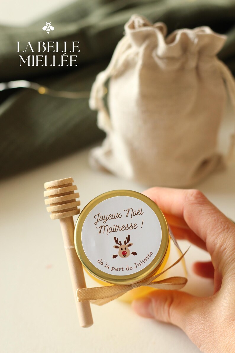 Pot de miel artisanal personnalisé avec étiquette 'Joyeux Noël Maîtresse', décoré d’un ruban beige et d’une cuillère en bois. Cadeau local et unique pour Noël, parfait pour remercier les maîtresses . Produit dans la Drôme