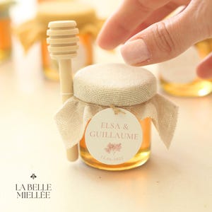 Peut inclure: Un petit pot en verre rempli de miel, surmonté d'un tissu en toile de jute et d'une étiquette ronde blanche sur laquelle est inscrit "Elsa & Guillaume 15.06.2025". Un bâtonnet à miel en bois est attaché au pot.