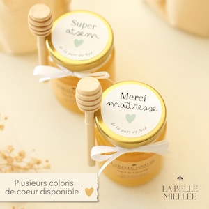 Peut inclure: Deux petits pots de miel avec des couvercles dorés et des cuillères à miel en bois. Les pots ont des étiquettes blanches avec du texte français et un cœur. Un ruban blanc est noué autour de chaque pot. Le texte "LA BELLE MIELLEE" est visible.
