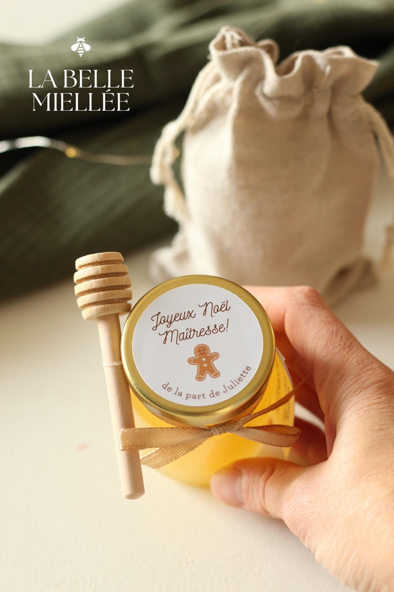 Petit pot de miel personnalisé pour Noël avec étiquette 'Joyeux Noël Maîtresse', cuillère en bois et ruban beige, idéal comme cadeau artisanal et local pour remercier une enseignante. Produit par La Belle Miellée dans la Drôme