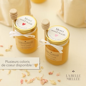 Peut inclure: Deux petits pots en verre remplis de miel doré, chacun avec un couvercle doré et une étiquette « Merci maîtresse ». Un bâtonnet à miel en bois repose sur chaque pot. Des rubans blancs et une petite pancarte avec du texte sont également visibles.