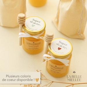 Peut inclure: Deux petits pots de miel en verre avec des cuillères à miel en bois. Les pots ont des couvercles dorés avec des étiquettes indiquant "Super atsem". Des rubans blancs sont noués autour des pots. Le texte "LA BELLE MIELLEE" est visible sur les étiquettes.