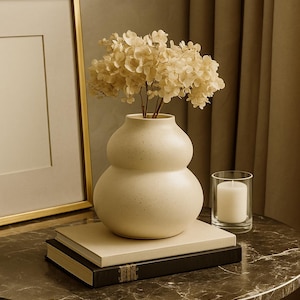 May include: A cream-colored, double-bulb vase holds dried flowers. The vase sits atop two books on a marble-topped table. A candle in a glass holder and a framed picture are also on the table.