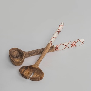 Puede incluir: Dos utensilios de ensalada de madera. Uno es una cuchara, el otro tiene un diseño ranurado. Los mangos son blancos con un patrón de lazo rojo. Las cucharas de madera son de color marrón claro.