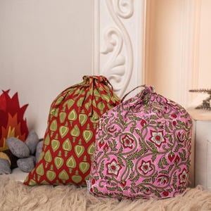 May include: Two drawstring bags with vibrant patterns. One is red with green leaf motifs, the other pink with a floral design. Both bags are tied at the top and rest on a fluffy, light-coloured surface.