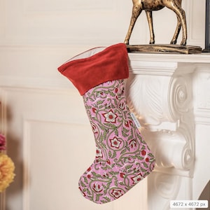 May include: A pink Christmas stocking with a floral design in red and green. The cuff is red, and the stocking hangs from a white mantelpiece. A gold deer statue is on the mantel.