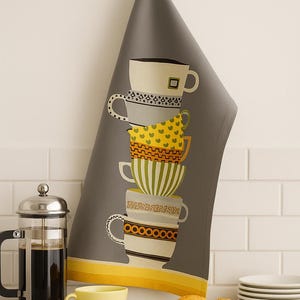 Puede incluir: Paño de cocina gris con una pila de tazas de café ilustradas en varios patrones y colores. Se ve un diseño de rayas amarillas y blancas, un patrón de corazones amarillos y un patrón geométrico en blanco y negro. Una taza amarilla y magdalenas están en el mostrador.