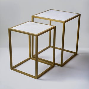 Puede incluir: Dos mesas auxiliares modernas. Cada mesa tiene una tapa cuadrada de mármol blanco y un marco de metal dorado. Las mesas tienen forma de cubo, con la mesa más pequeña dentro de la más grande. Sobre un fondo blanco.