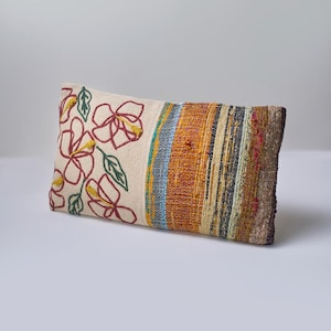 May include: A long rectangular pillow with a white fabric cover featuring embroidered red flowers and green leaves. The back of the pillow is made of a woven fabric with colorful stripes in shades of orange, yellow, blue, and brown.