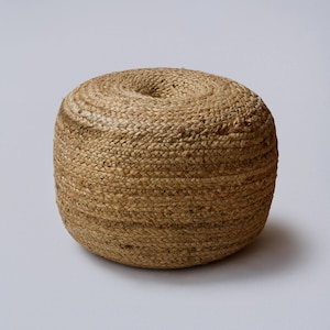 Op de afbeelding: Een ronde, geweven jute poef in natuurlijke tinten. De poef heeft een cilindrische vorm met een licht ingedeukte bovenkant. De textuur is grof en de kleur is een warm, aards bruin. Dit decoratieve stuk is geschikt voor binnengebruik.