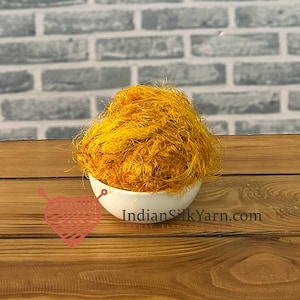 May include: A mound of vibrant golden-yellow textured silk yarn fibers contained within a small white ceramic bowl. The text IndianSilkYarn.com is printed on the side of the bowl, accompanied by a red graphic of a heart-shaped ball of yarn.
