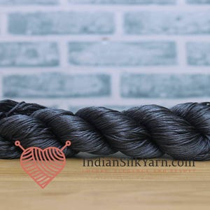 May include: A skein of dark grey silk yarn, tightly twisted and displayed on a wooden surface. The yarn has a subtle sheen, reflecting light. The background features a light blue brick wall. The logo of IndianSilkYarn.com is visible.
