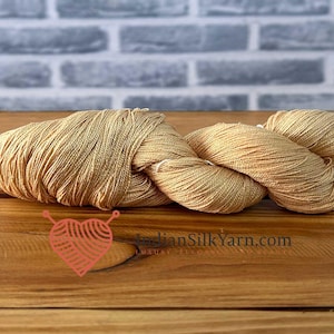 Op de afbeelding: Een streng goudkleurig zijdegaren, gepresenteerd op een houten oppervlak. Het garen is strak gewikkeld en ziet er zacht en luxueus uit. De website "IndianSilkYarn.com" is zichtbaar in de afbeelding.