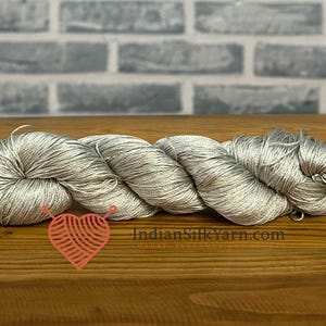 Puede incluir: Una madeja de hilo plateado brillante con una textura suave y lustrosa. El hilo está retorcido en una bobina larga y continua. Un logotipo rojo en forma de corazón con un gráfico de ovillo y el texto IndianSilkYarn.com es visible.